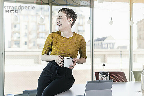 Lachende Geschäftsfrau  die auf einem Schreibtisch im Büro sitzt und einen Kaffeebecher hält