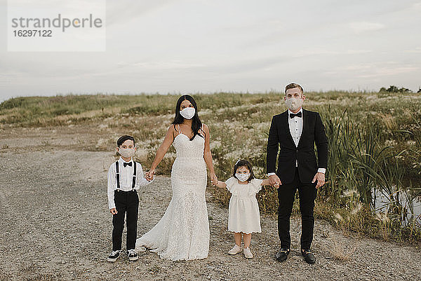 Eltern und Kinder in Hochzeitskleidern tragen einen Mundschutz  während sie während COVID-19 im Feld stehen