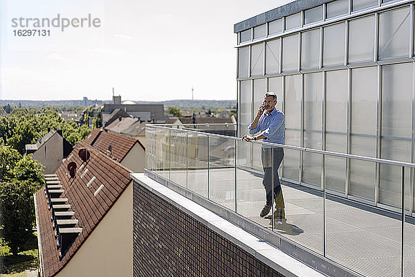 Geschäftsmann im Gespräch über Smartphone  während er auf dem Balkon gegen den Himmel an einem sonnigen Tag steht