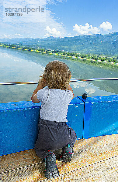 Kleiner Junge kniet vor einem Geländer und betrachtet die malerische Aussicht auf den Kerkini-See  Mazedonien  Griechenland