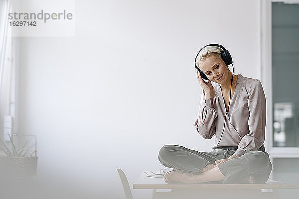 Weibliche Fachkraft hört Musik über Kopfhörer  während sie auf dem Schreibtisch an der Wand im Büro sitzt