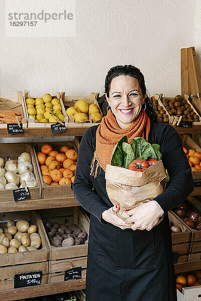 Gut gelaunte Verkäuferin  die eine Tüte mit Gemüse in der Hand hält  die vor Kisten im Laden steht