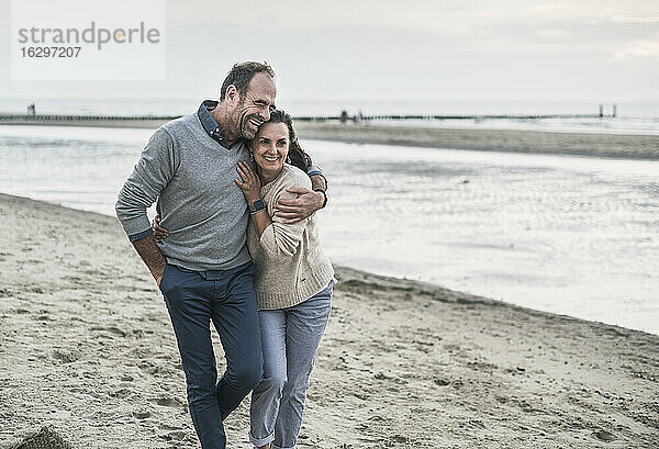 Fröhlicher Mann umarmt Frau beim Spaziergang am Strand gegen den Himmel