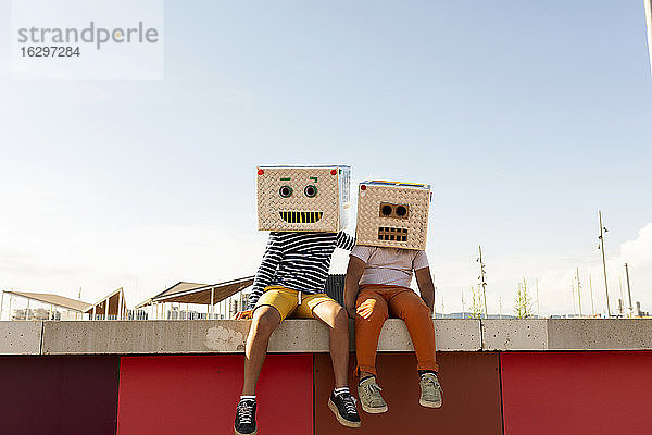 Freunde in Roboterkostümen sitzen im Sommer auf einer Stützmauer gegen den klaren Himmel