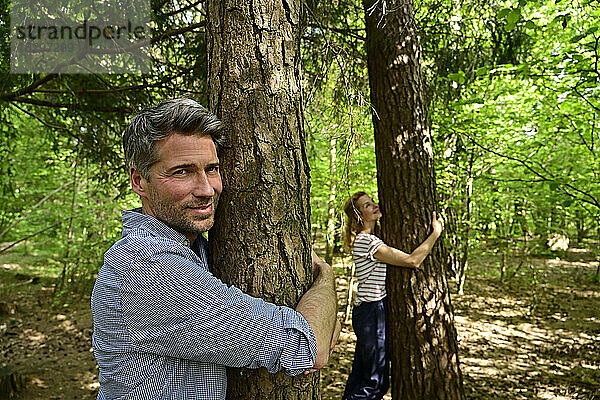 Lächelnder Mann und lächelnde Frau umarmen einen Baum  während sie im Wald stehen