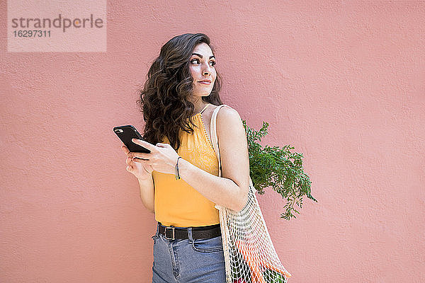 Junge Frau blickt zurück  während sie ihr Smartphone hält und mit einer wiederverwendbaren Netztasche vor einer rosafarbenen Wand steht