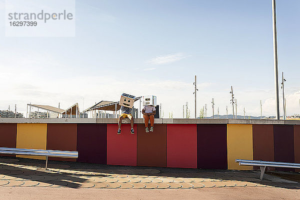 Freunde in Roboterkostümen sitzen auf einer bunten Stützmauer gegen den Himmel an einem sonnigen Tag