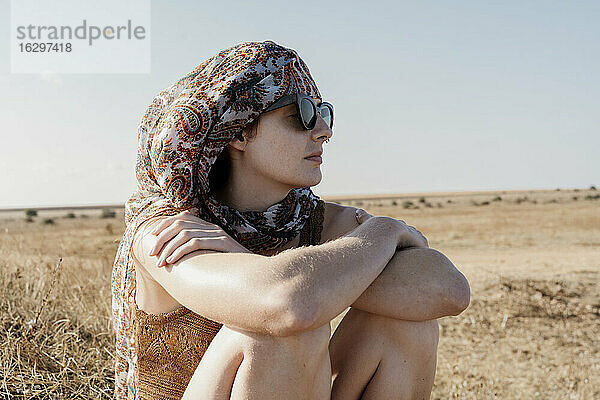 Porträt einer jungen Frau mit Kopftuch und Sonnenbrille  die mit verschränkten Armen im Freien sitzt