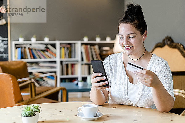 Lächelnde Frau mit Kaffee auf dem Tisch beim Online-Shopping mit dem Handy im Café