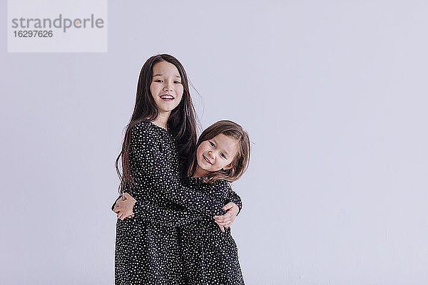 Lächelnde Schwestern  die sich umarmen  während sie vor einem weißen Hintergrund im Studio stehen