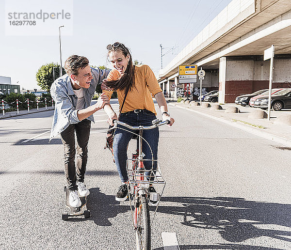 Verspielter Freund auf dem Skateboard  während seine Freundin auf der Straße in der Stadt Fahrrad fährt
