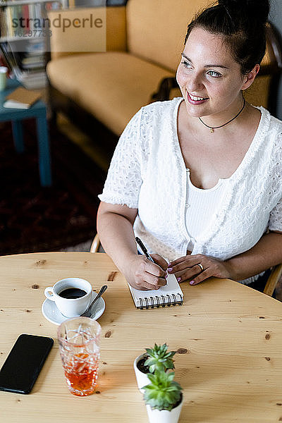 Junge Frau  die nachdenklich in einen Notizblock auf einem Tisch in einem Café schreibt