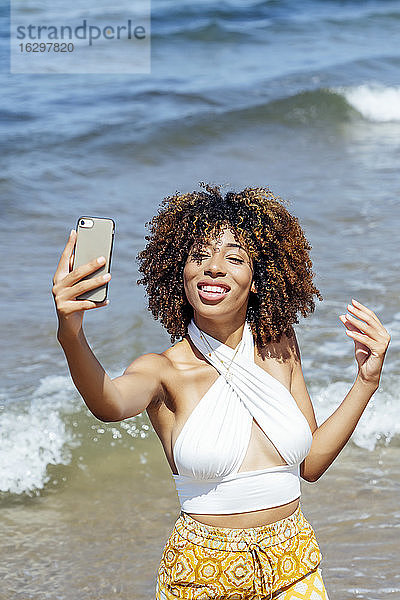 Junge Frau mit lockigem Haar  die ein Selfie macht  während sie an einem sonnigen Tag im Meer steht