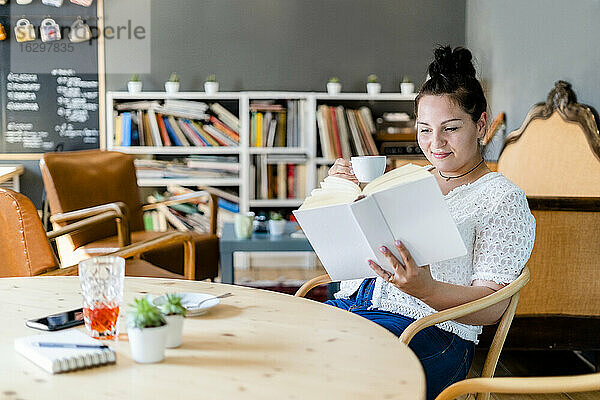 Junge Frau  die eine Kaffeetasse hält und ein Buch liest  während sie am Tisch in einem Café sitzt
