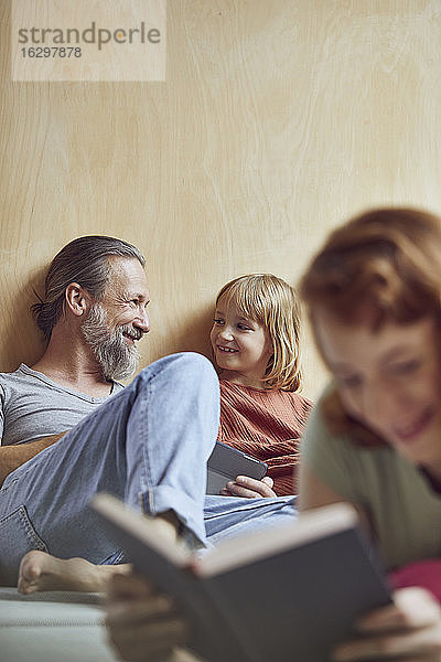 Lächelnder Vater und Tochter schauen sich an  während die Frau zu Hause auf dem Bett ein Buch liest