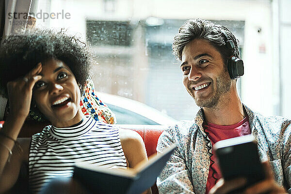 Lächelnder Mann  der Musik hört  während seine Freundin mit einem Buch am Fenster eines Cafés sitzt