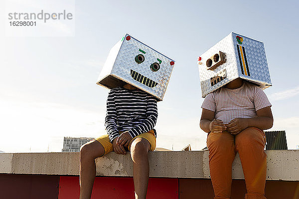 Freunde mit Robotermasken sitzen auf einer Stützmauer gegen den klaren Himmel an einem sonnigen Tag