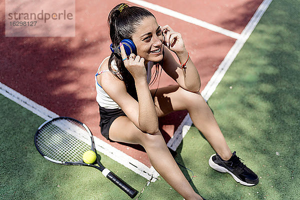 Lächelnde Tennisspielerin hört Musik  während sie auf dem Boden im Gericht sitzt