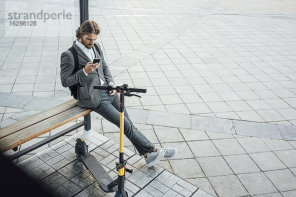 Männlicher Unternehmer  der ein Mobiltelefon benutzt  während er auf einer Bank mit einem Elektroroller in der Innenstadt sitzt