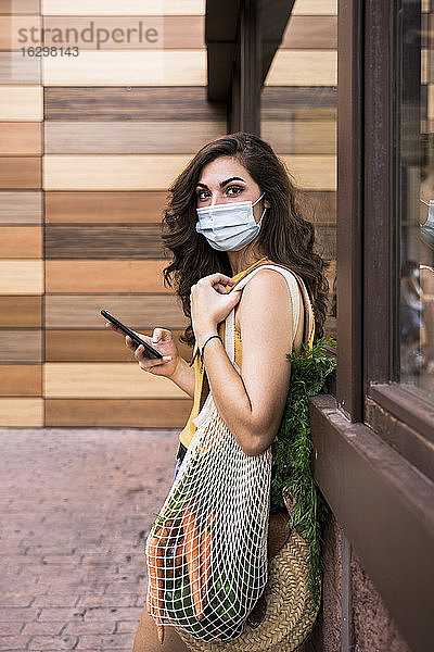 Junge Frau mit Schutzmaske  die ein Mobiltelefon und eine Netztasche hält  während sie an der Wand steht
