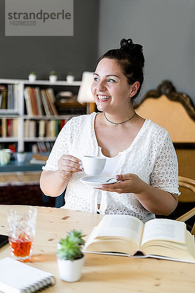 Glückliche junge Frau mit Buch auf dem Tisch  die eine Kaffeetasse hält  während sie im Restaurant sitzt