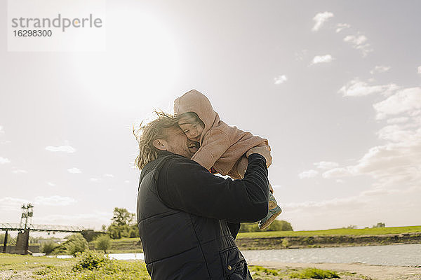 Vater kuschelt mit seiner Tochter  während er an einem sonnigen Tag am Flussufer steht