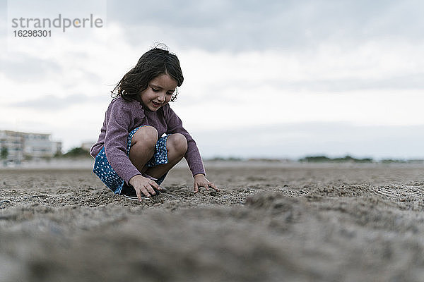 Lächelndes Mädchen  das mit Sand spielt  während es am Strand vor einem bewölkten Himmel kauert