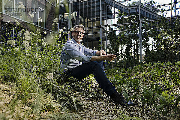 Nachdenklicher Geschäftsmann schaut weg  während er inmitten von Pflanzen sitzt