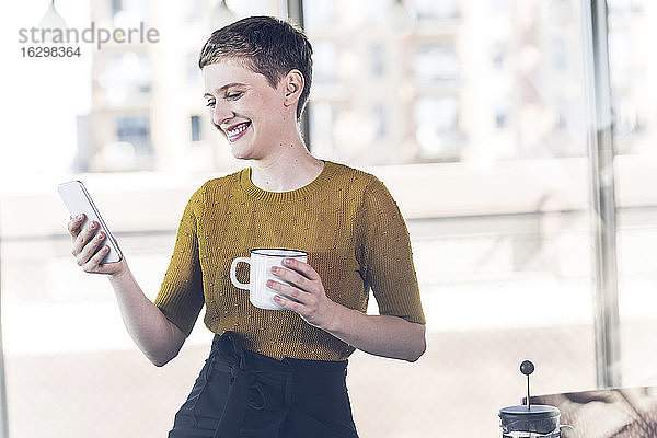 Lächelnde Geschäftsfrau im Büro mit Kaffeetasse und Mobiltelefon