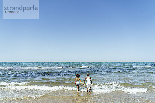 Paar hält Hände beim Spaziergang im Meer gegen den klaren Himmel