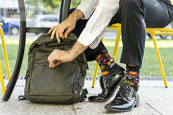 Männlicher Unternehmer mit bunten Socken  der seinen Rucksack schließt  während er auf einem Stuhl in einem Straßencafé sitzt