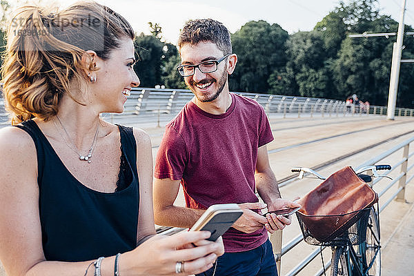 Glückliches Paar benutzt Smartphones auf einer Brücke bei Sonnenuntergang