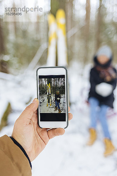 Freund fotografiert Freundin mit Skiern durch Handy im Wald im Winter