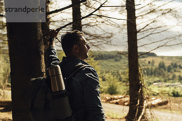 Nachdenklicher männlicher Wanderer mit Rucksack an einem Baumstamm im Wald stehend
