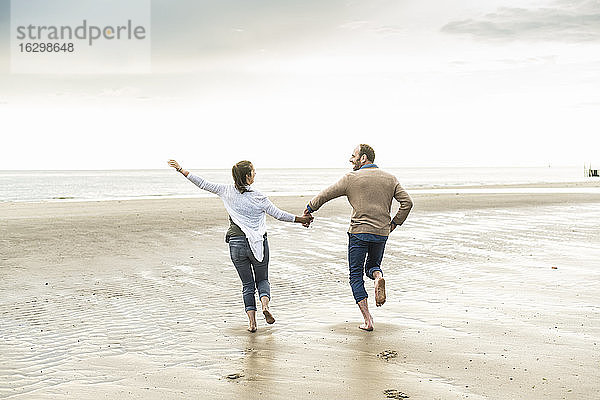 Fröhliches Paar hält sich beim Laufen am Strand während des Sonnenuntergangs an den Händen