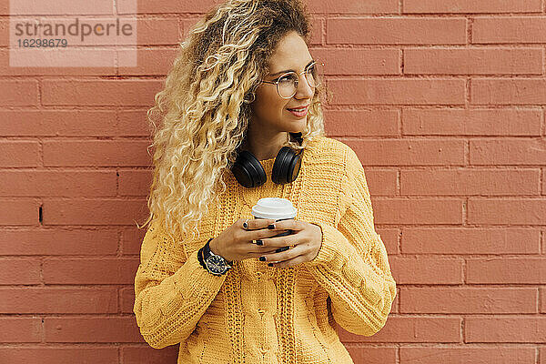 Junge Frau mit blondem  lockigem Haar  die wegschaut  während sie eine Einweg-Kaffeetasse gegen eine rote Backsteinwand hält