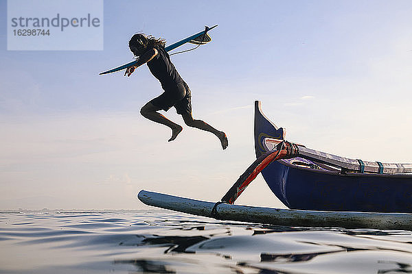 Mann mit Surfbrett springt in Meer gegen klaren blauen Himmel