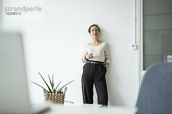Selbstbewusste Geschäftsfrau mit Hand in der Tasche  die eine Kaffeetasse hält  während sie vor einer weißen Wand steht
