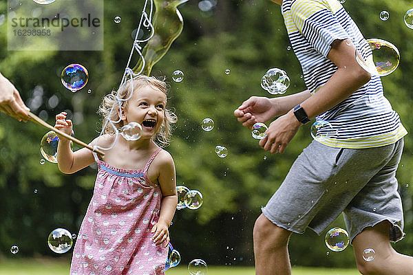 Fröhliches Mädchen vergnügt sich mit seinem Bruder beim Laufen inmitten von Seifenblasen im öffentlichen Park