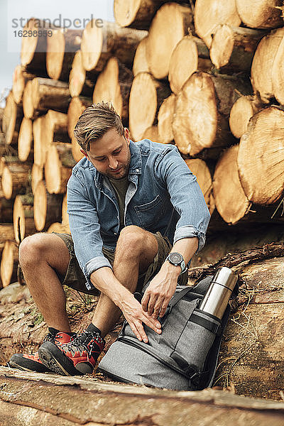 Männlicher Wanderer mit Rucksack auf einem Baumstamm sitzend gegen einen Holzstapel im Wald