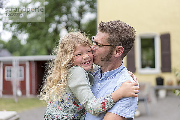 Mann küsst glückliche blonde Tochter  während er sie im Hinterhof trägt