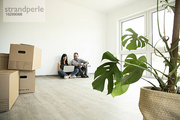 Mann sitzt mit Frau vor Laptop auf Hartholzboden in einem neuen unmöblierten Haus