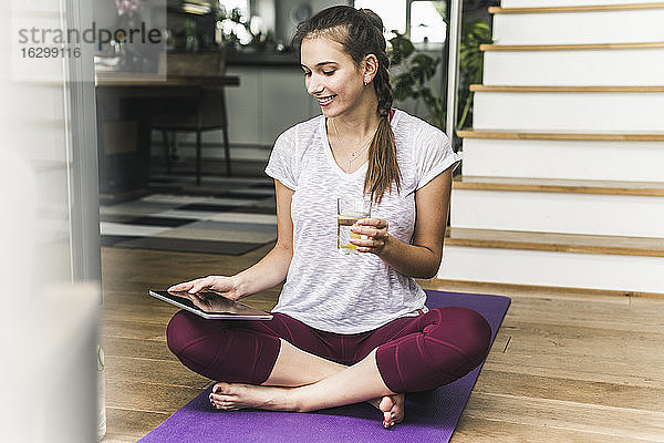 Lächelnde Frau hält ein Glas  während sie ein digitales Tablet auf einer Übungsmatte zu Hause benutzt