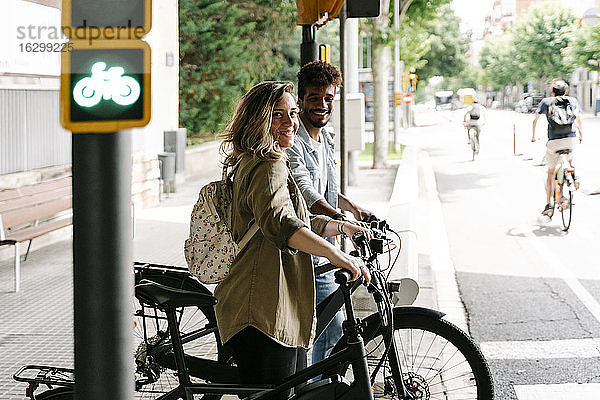 Lächelndes junges Paar mit Elektrofahrrädern auf dem Bürgersteig stehend