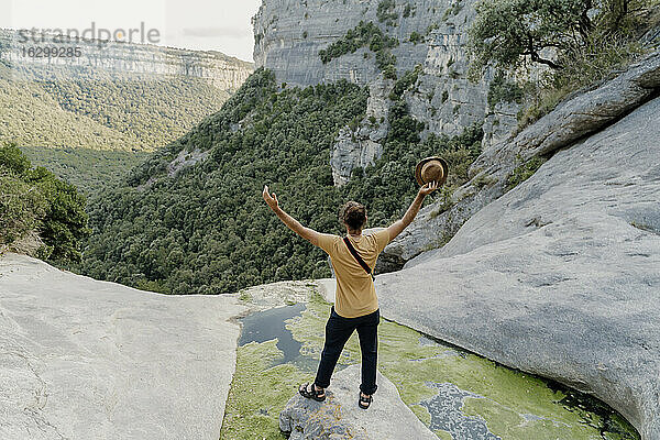 Männlicher Wanderer steht mit erhobenen Armen auf einem Felsen inmitten eines Gebirgsbachs mit einem bewaldeten Tal im Hintergrund