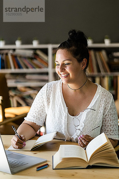 Lächelnde junge Frau  die in ein Buch schreibt  während sie über einem Laptop auf dem Tisch eines Cafés studiert