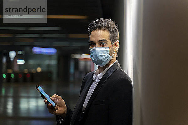 Geschäftsmann mit Maske  der ein Smartphone benutzt  während er an der Wand eines Bahnhofs steht