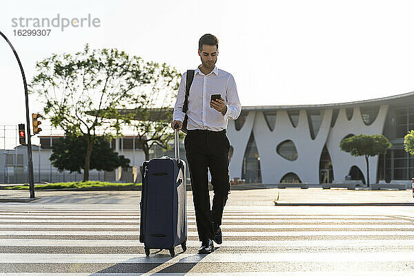 Geschäftsmann mit Koffer und Smartphone beim Überqueren der Straße gegen den klaren Himmel