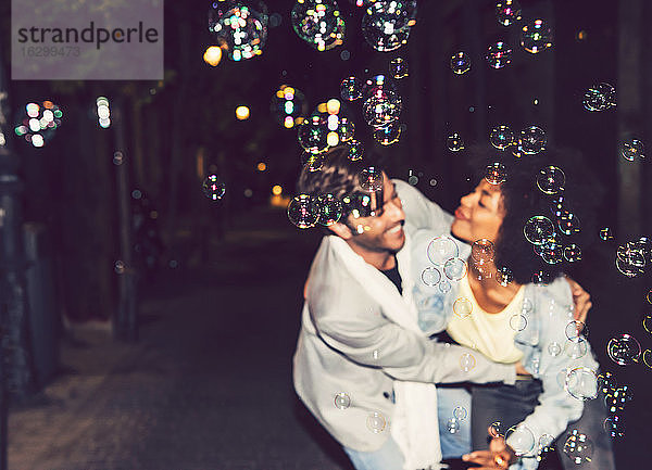 Nahaufnahme von Seifenblasen mit tanzendem Paar auf der Straße im Hintergrund bei Nacht