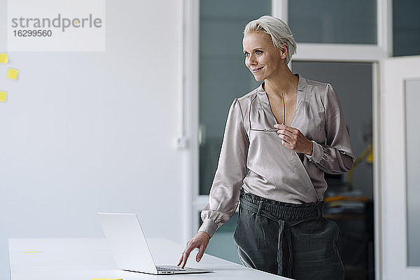 Lächelnde weibliche Fachkraft  die im Büro stehend einen Laptop auf dem Schreibtisch benutzt